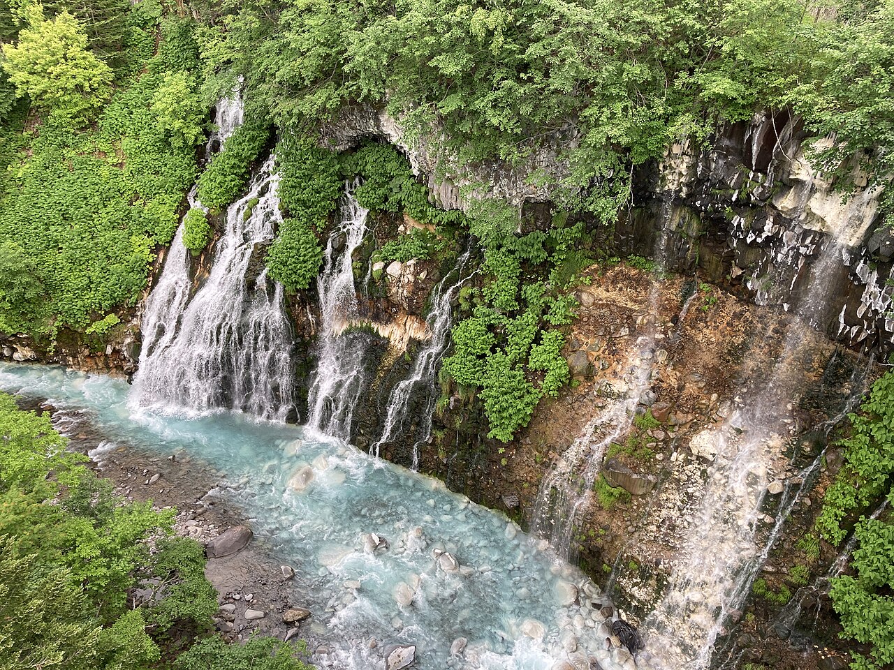 Shirahige Falls pouring from a green cliff face into the pale blue Biei River