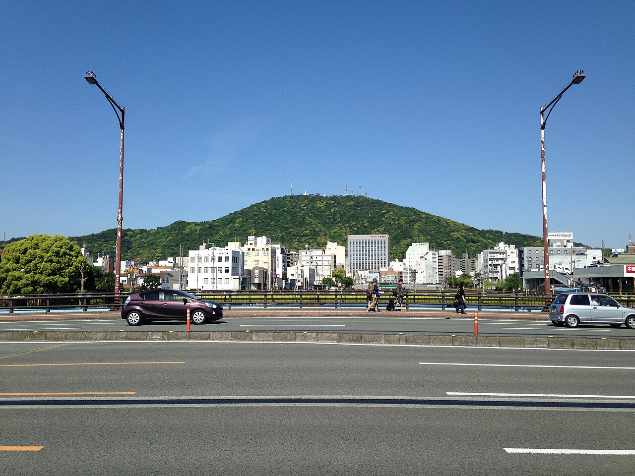 The low green mass of Mount Bizan rising behind city buildings, seen across a broad road and bridge on a clear day