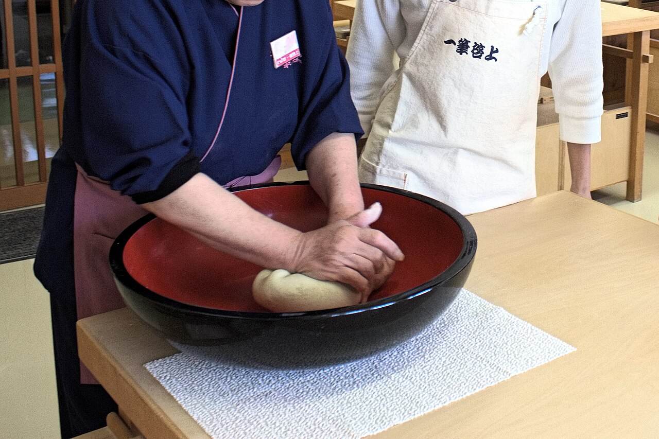 Hands kneading buckwheat dough in a large red lacquered bowl at a soba-making table, with a person in an apron standing behind