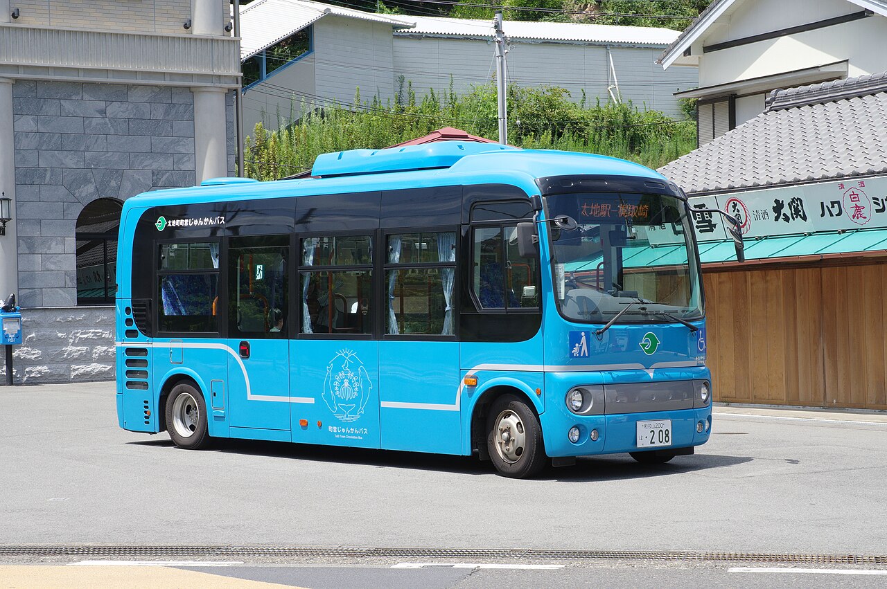 A small light-blue town circulation bus stopped on a quiet street in front of old shop buildings