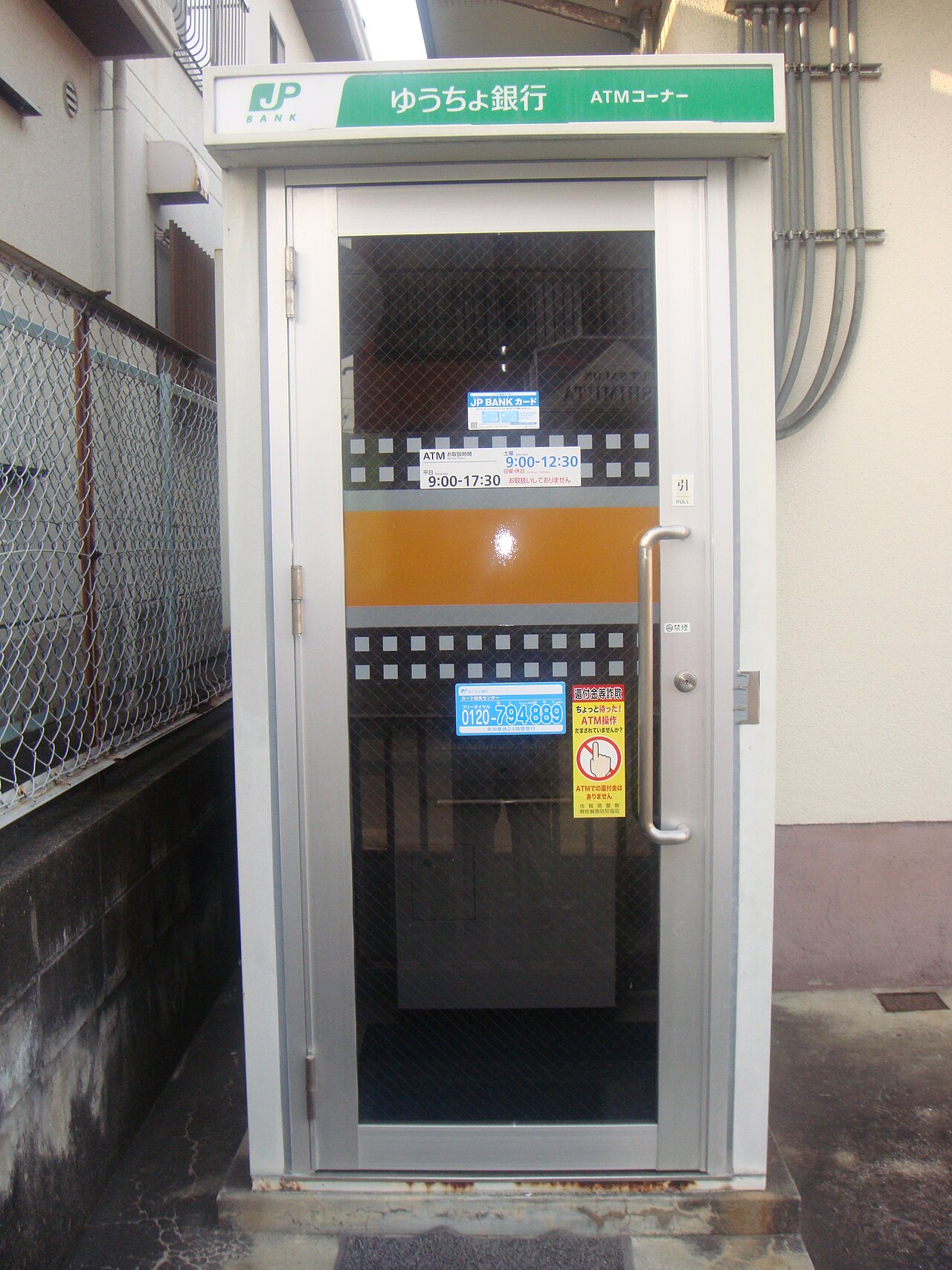 A small freestanding Japan Post Bank ATM booth on a residential street, with the green JP Bank ATM corner sign above a glass door