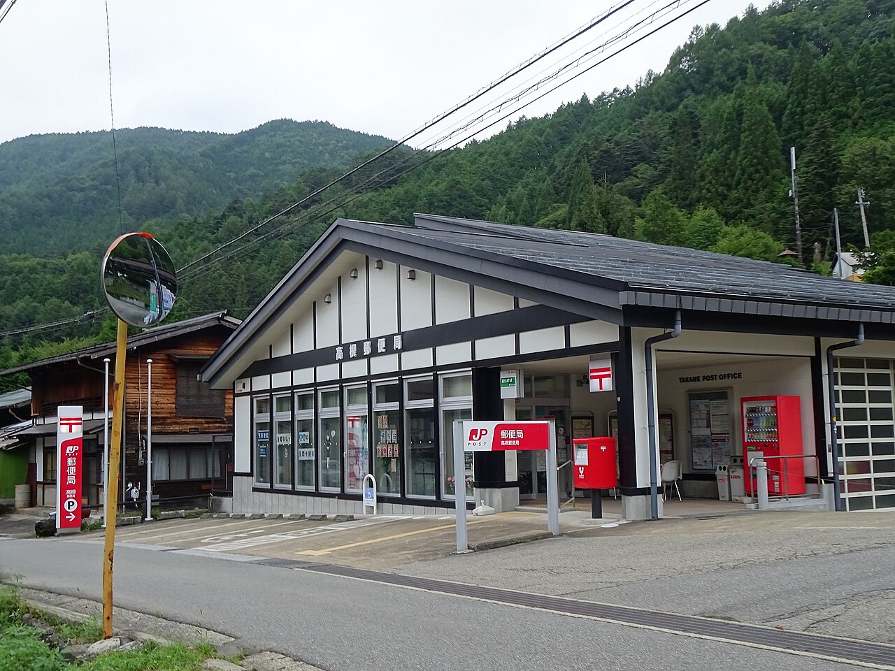 Takane post office in a mountain village, with a red Japan Post sign, a red mailbox and a vending machine under the eaves and forested hills behind