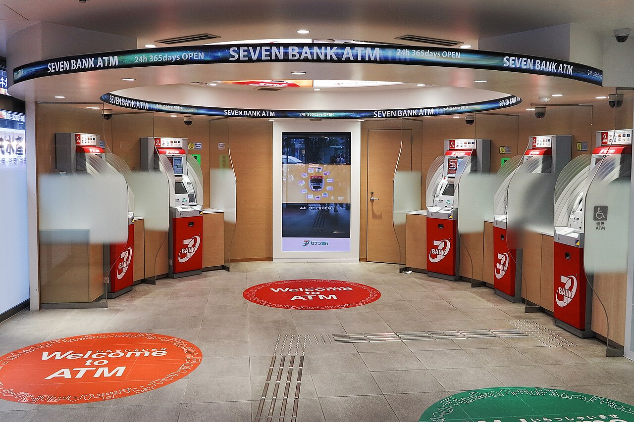 A row of red Seven Bank ATMs under a ring-shaped Seven Bank ATM sign, with Welcome to ATM stickers on the floor