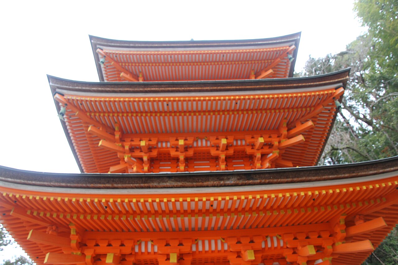 Looking up at the vermilion three-storied pagoda at Hogon-ji, its tiered eaves and bracket work against a pale sky
