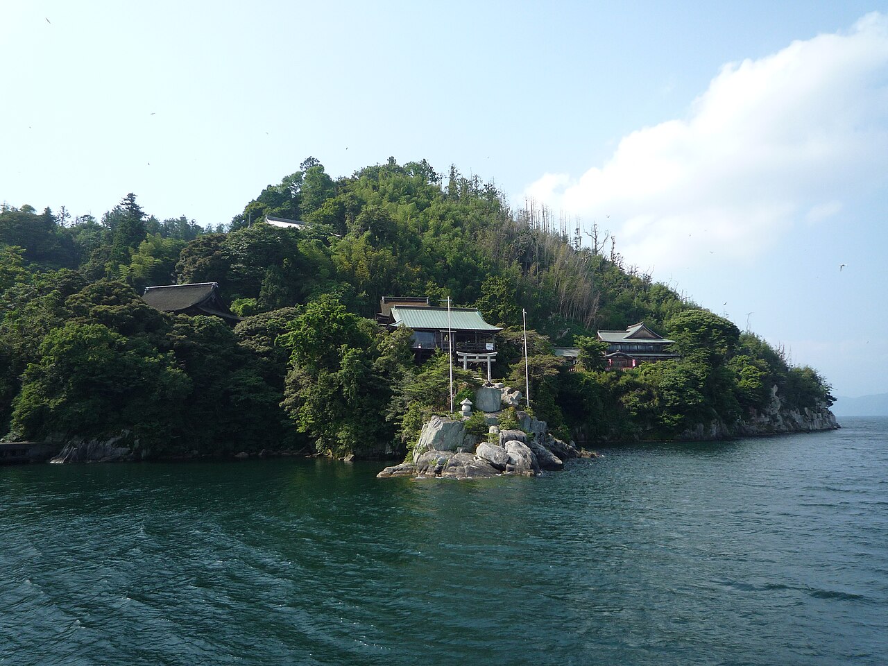 Wooded shoreline of Chikubushima seen from a boat on Lake Biwa, with a small torii on a rock at the water and shrine roofs among the trees