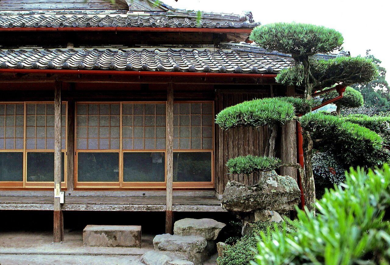 A close view of a tiled roof eave and a cloud-pruned pine rising beside a moss-covered boulder in a Chiran samurai garden.