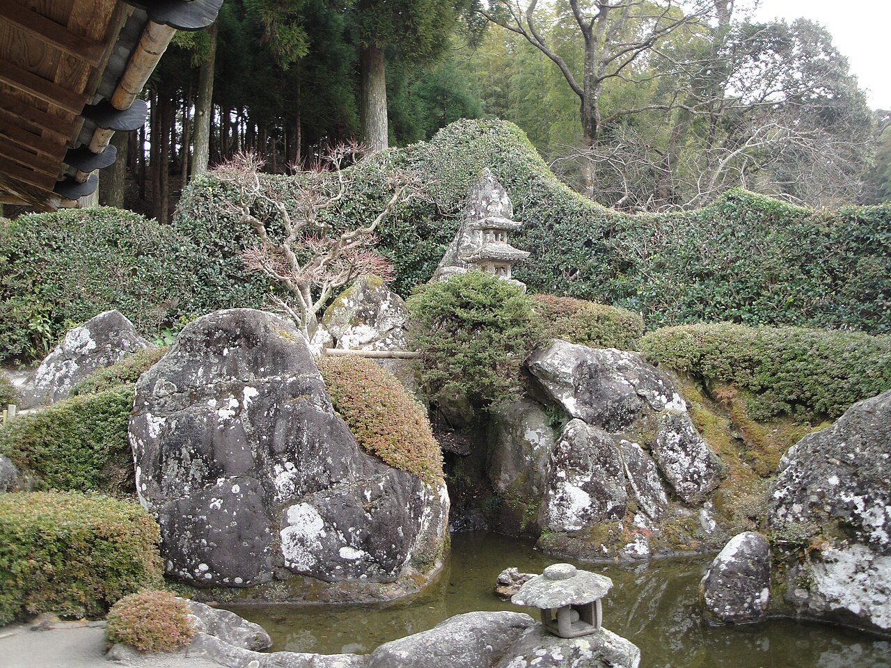 A koi pond edged with moss-covered boulders and a stone lantern in a Chiran samurai garden, framed by clipped hedges and cedar trees.