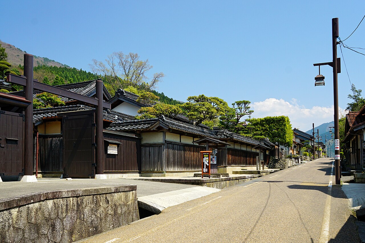 Dark timber gate with a torii-style crossbeam beside the white-and-dark-wood walls of the Chizu post town street, with wooded hills behind