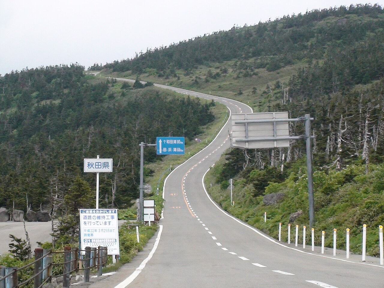 A two-lane mountain road climbing a forested ridge on the Hachimantai Aspite Line, with an Akita prefecture sign at the roadside