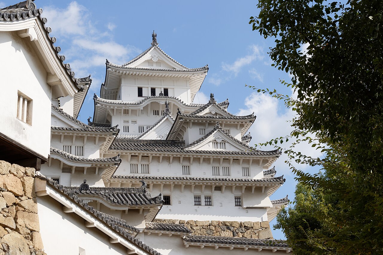The white tiered keep of Himeji Castle seen above stone walls and tiled roofs against a blue sky