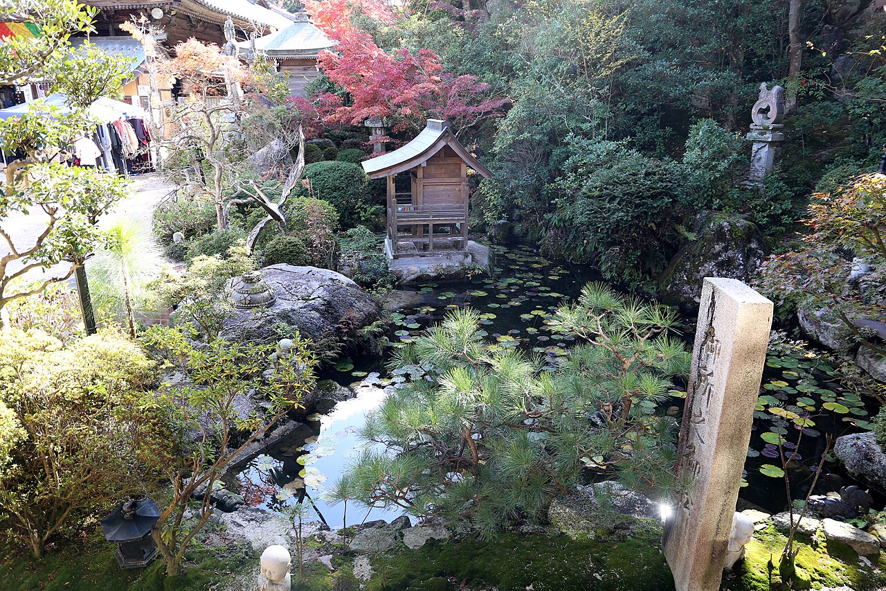A temple garden with a pond edged by rocks and clipped pines, a small wooden shrine among trees and a red maple leaf in the corner, with a stone marker in the foreground.