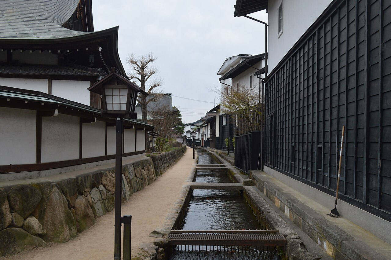 The narrow Setogawa canal in Hida Furukawa, with a white temple wall on a stone base to the left, a tall dark timber storehouse to the right, and lantern-shaped streetlights along the lane under a grey sky