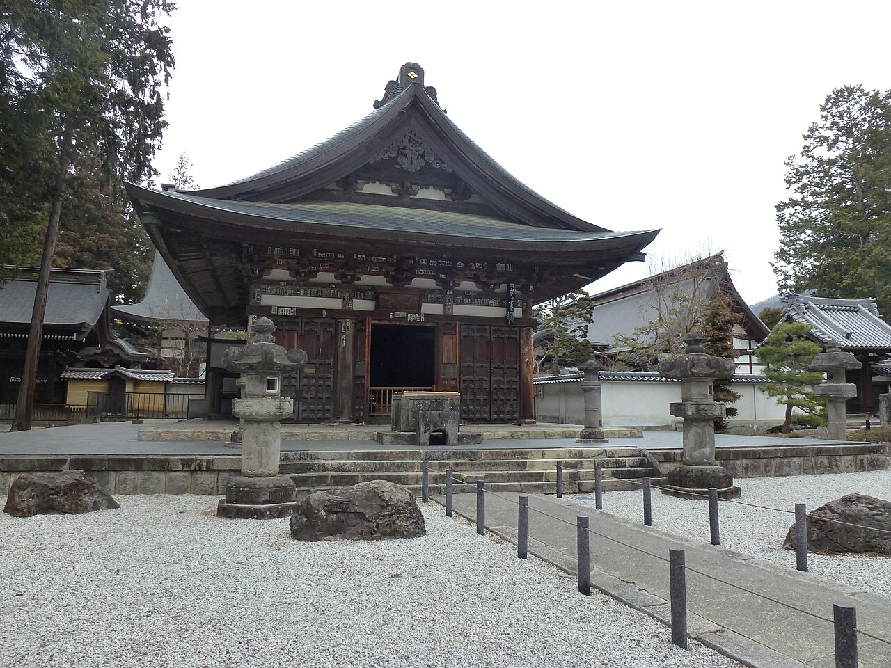 A wooden temple hall with a sweeping curved tiled roof standing behind a white gravel yard with stone lanterns and boulders, a rope fence along the path in front