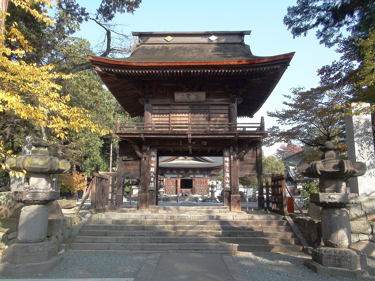 The two-storey Sanmon gate of Erinji seen from the stone steps, with stone lanterns on both sides, autumn leaves at the left and the main hall visible through the open passage