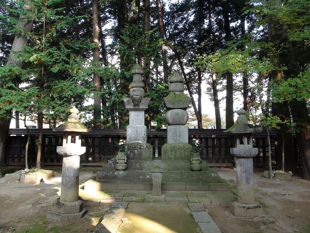 Two old stone pagodas on a stepped base at the Takeda Shingen grave site in Erinji, flanked by stone lanterns with offerings of flowers and tall cedars behind a dark wooden fence
