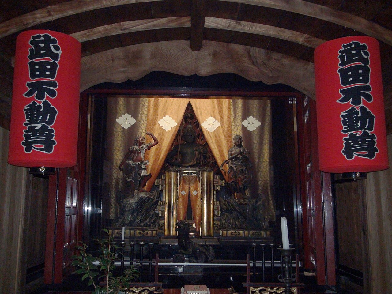 The Takeda Fudo statue enshrined in a small hall, framed by two red lanterns reading Takeda Fudoson and orange curtains with gold crests