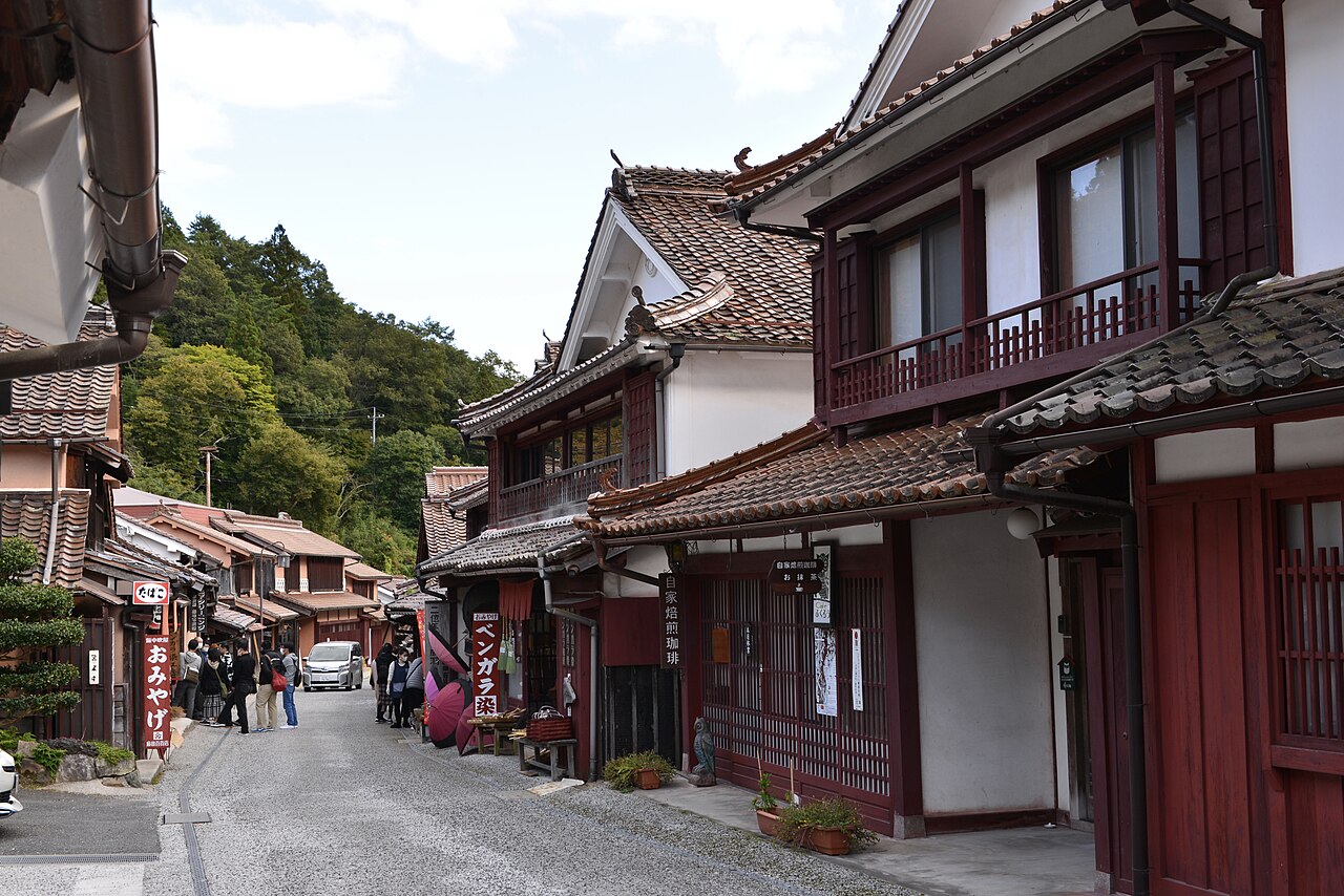 Red-lacquered lattice shopfronts and tiled roofs along Fukiya's stone-paved lane, with a few visitors walking in the distance
