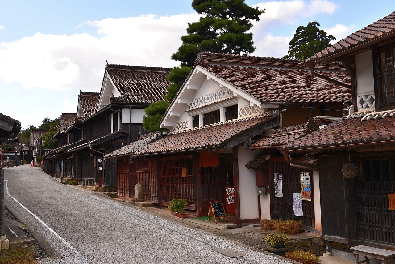 A gently sloping gravel street in Fukiya lined with old merchant houses with dark lattice windows, red-brown tiled roofs and a pine tree behind