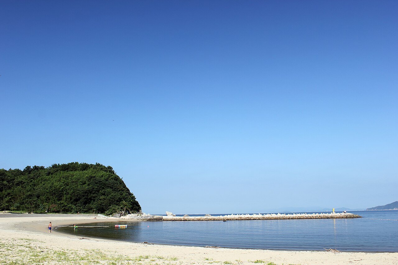 A curved sand beach on Nushima with a breakwater and a wooded headland under a clear sky
