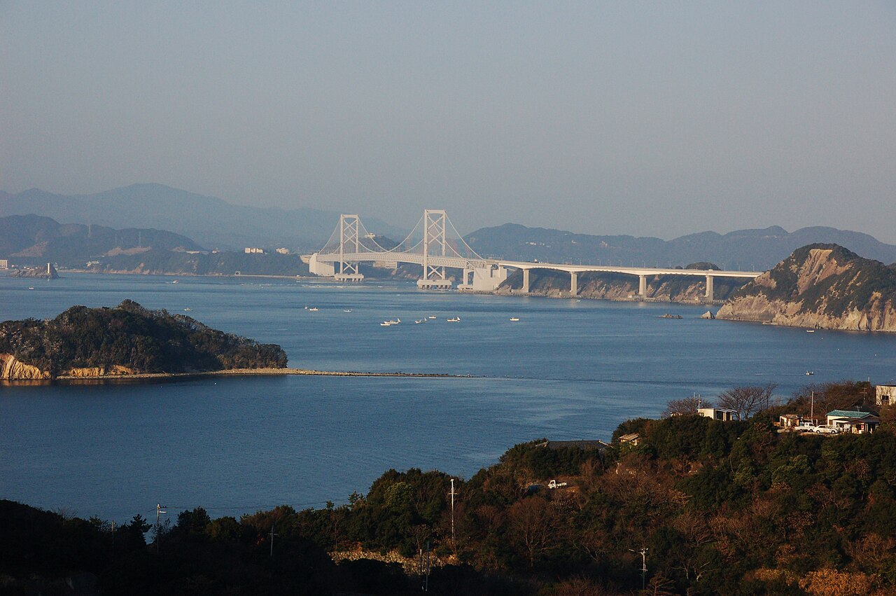 The Onaruto Bridge spanning the Naruto Strait beyond a wooded headland and a small island, with fishing boats on calm blue water