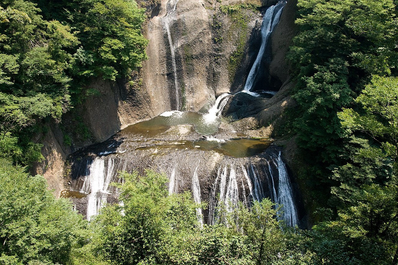 Looking down on the falls in summer: an upper cascade spills into a rock pool, then fans out into many white streams across a wide lower ledge, framed by green trees.