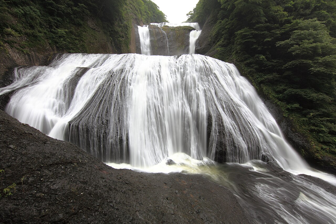 A wide, broad curtain of white water spreading down a rock face in a forested gorge, with the upper tiers of the falls visible between the dark green slopes.