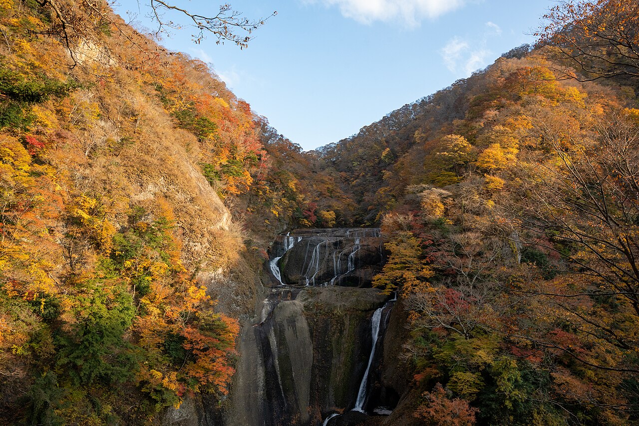 A steep gorge with orange and red autumn foliage on both slopes and a multi-tiered waterfall dropping at the far end under a blue sky.