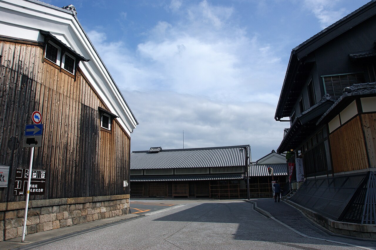 A quiet street between old sake-brewery buildings, with a dark wooden storehouse wall with white-plaster trim on the left, a low tiled-roof brewery building ahead and a wooden townhouse on the right under a blue sky with clouds.