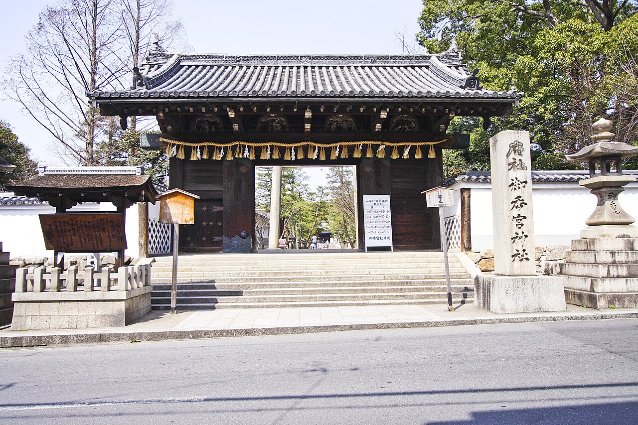 A dark wooden shrine gate with a heavy tiled roof and a twisted straw rope across its front stands at the top of wide stone steps, beside a tall stone marker engraved with the shrine name and a stone lantern, with green trees behind.