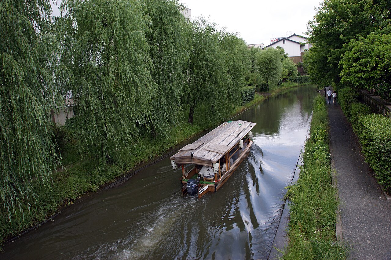 A wooden canal boat with paper lanterns glides down a narrow green canal lined with drooping willow trees, with a gravel towpath on the right and white-walled buildings behind.