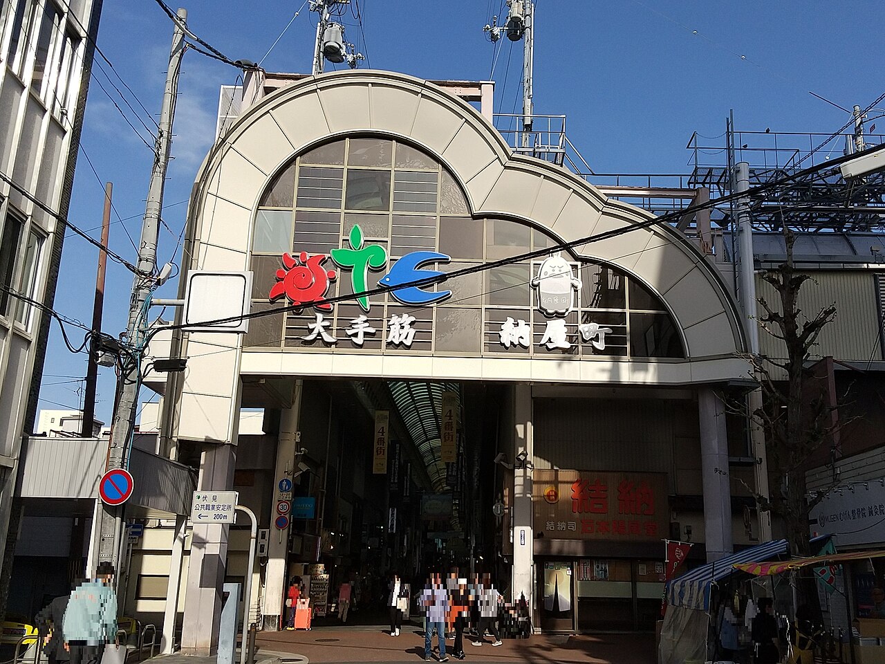 A broad arched entrance of a covered shopping street with colourful logo lettering on its glass facade, leading into a long arcade, with shoppers walking beneath and power lines crossing overhead.