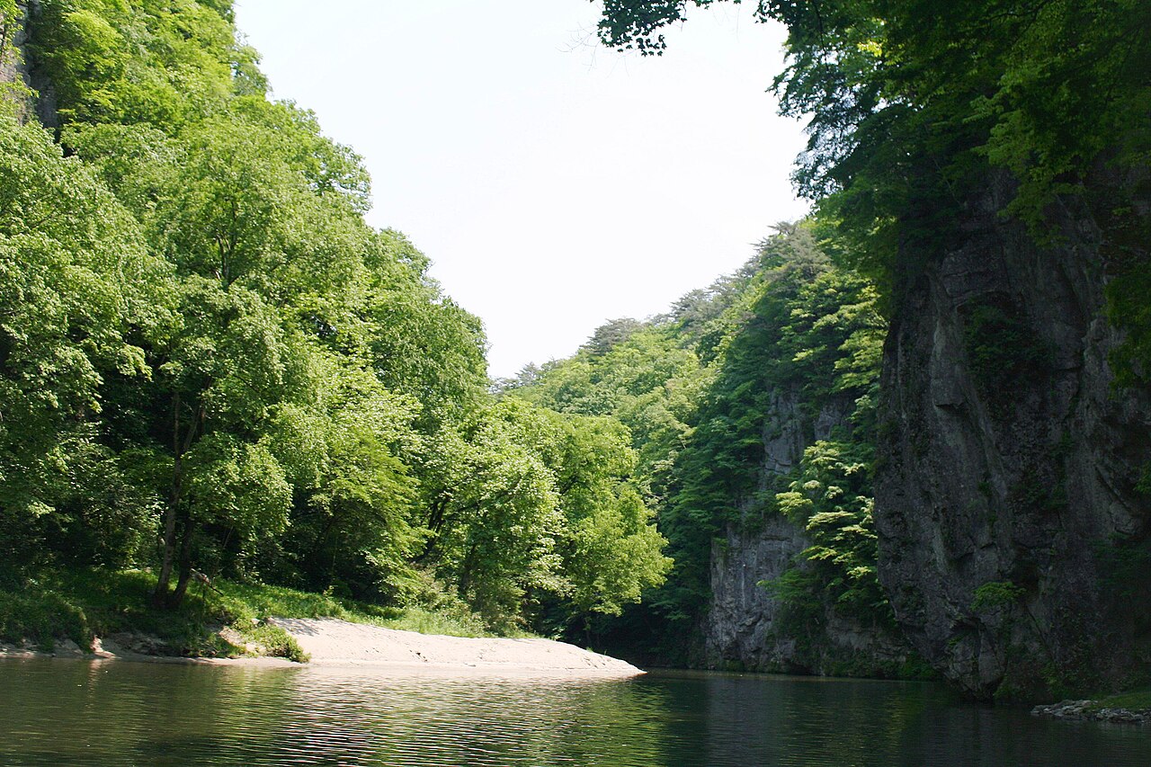 The Satetsu River bending through Geibikei Gorge between a steep rock wall on the right and bright green trees on the left, with a pale gravel bar in the middle