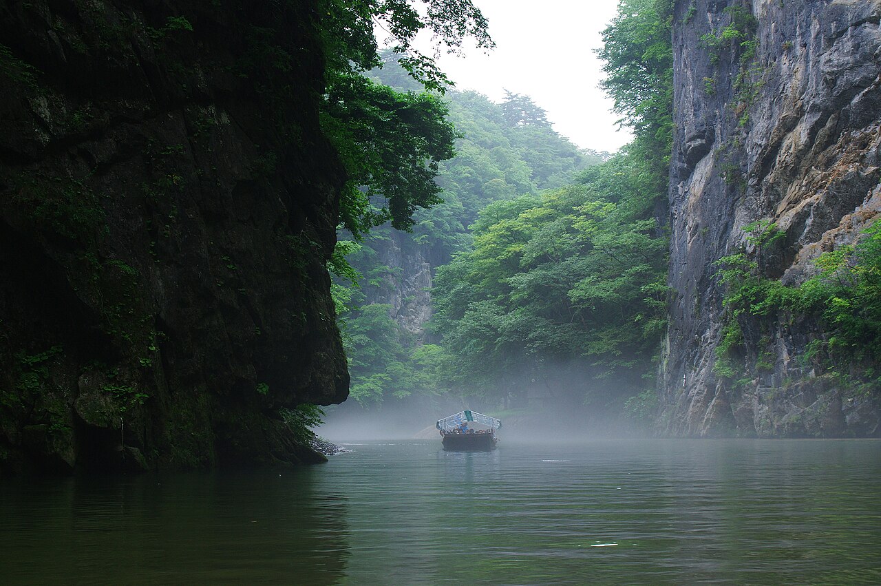 A small punt on the misty green water of the Satetsu River, with dark limestone cliffs and forested slopes on both sides of Geibikei Gorge