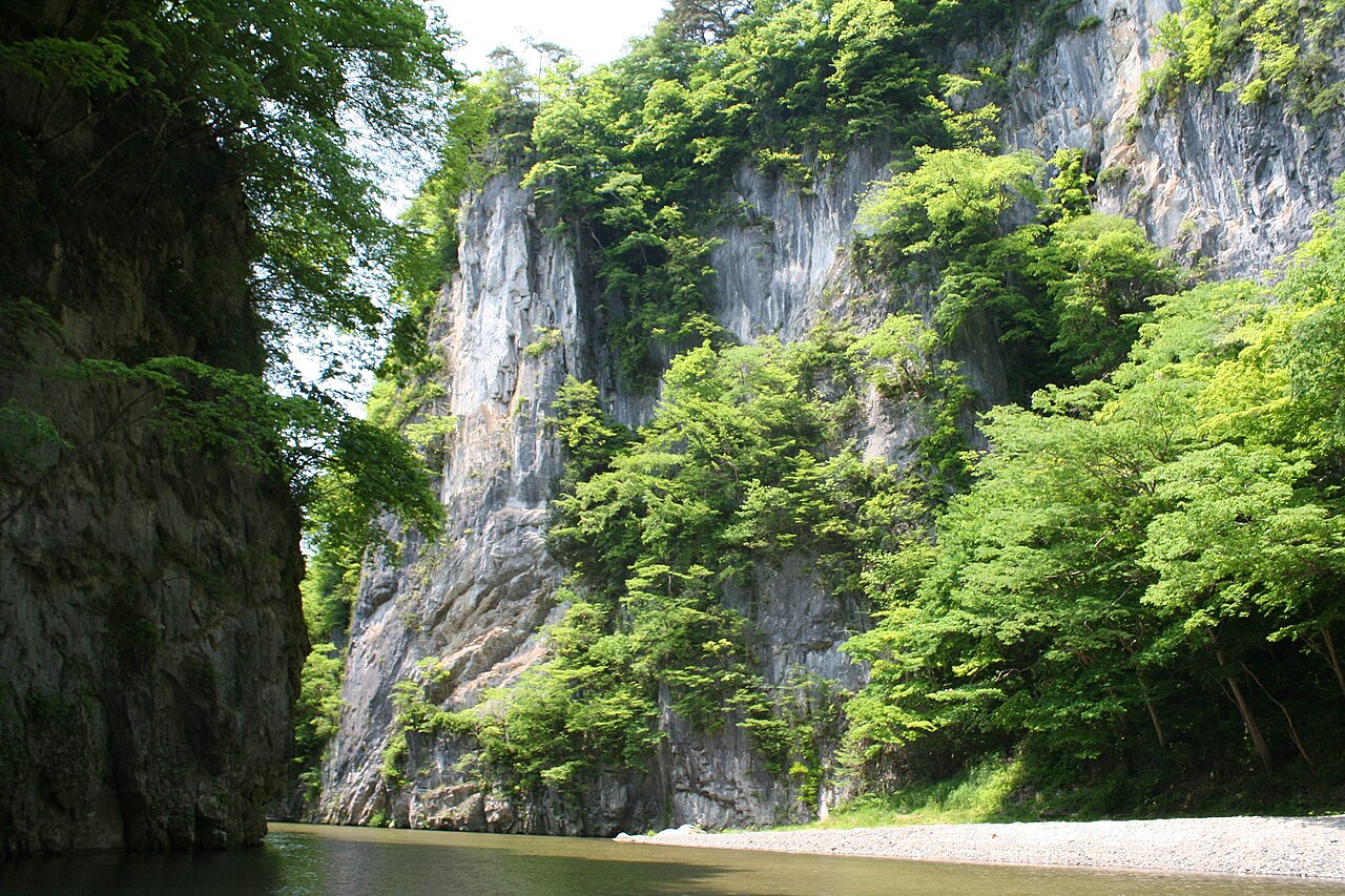 A tall pale limestone pillar draped in green trees beside dark rock, with a pebbly river bank at its foot in Geibikei Gorge