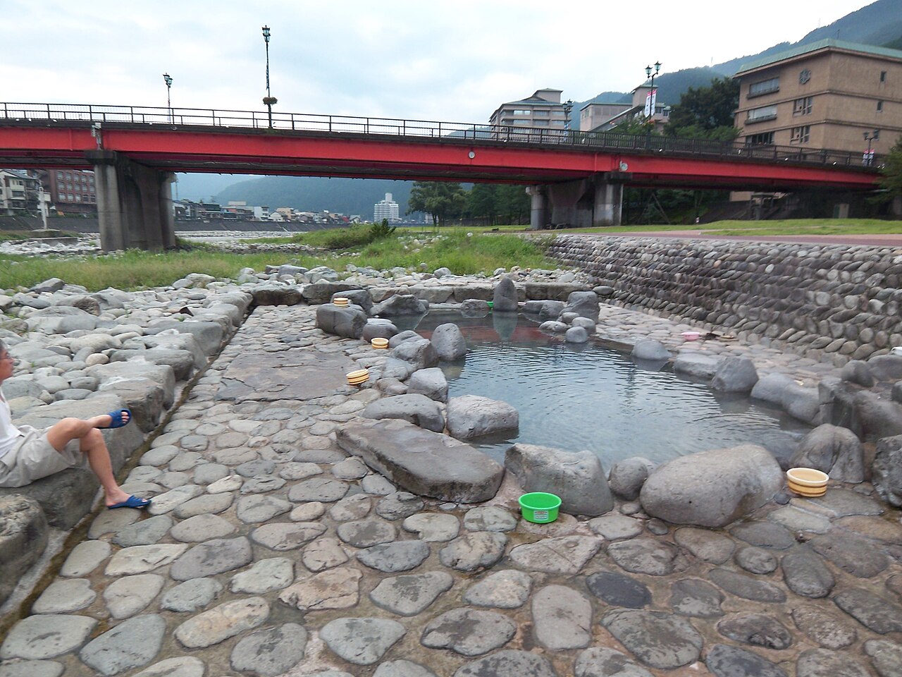 A stone-lined hot spring pool on the Hida River bank at Gero Onsen with a red bridge and the town behind it