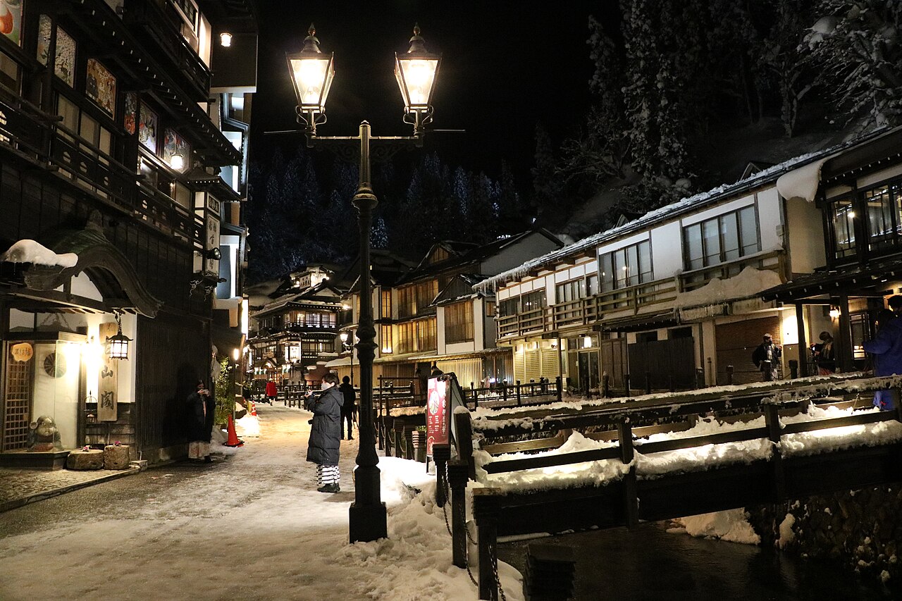 A gas lamp glowing on a snowy night street in an onsen town, with wooden inns and a bridge in the background.