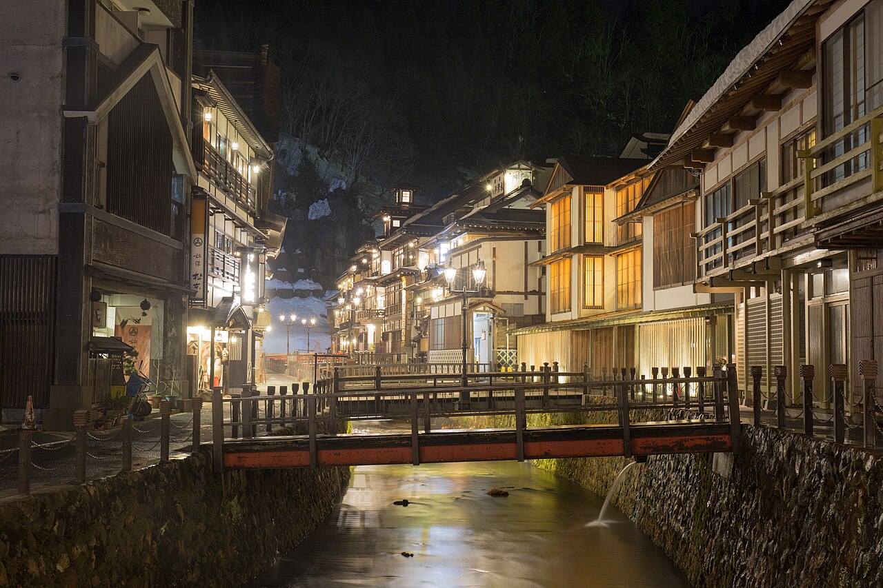 A red wooden bridge over a narrow river at night, with lit wooden inns lining the banks of the onsen town.