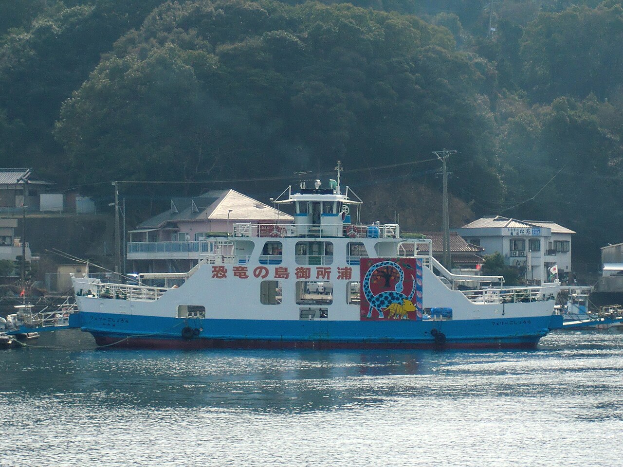 The blue-and-white car ferry Goshoura sits in the harbor under green hills, with a dinosaur picture painted on its side.