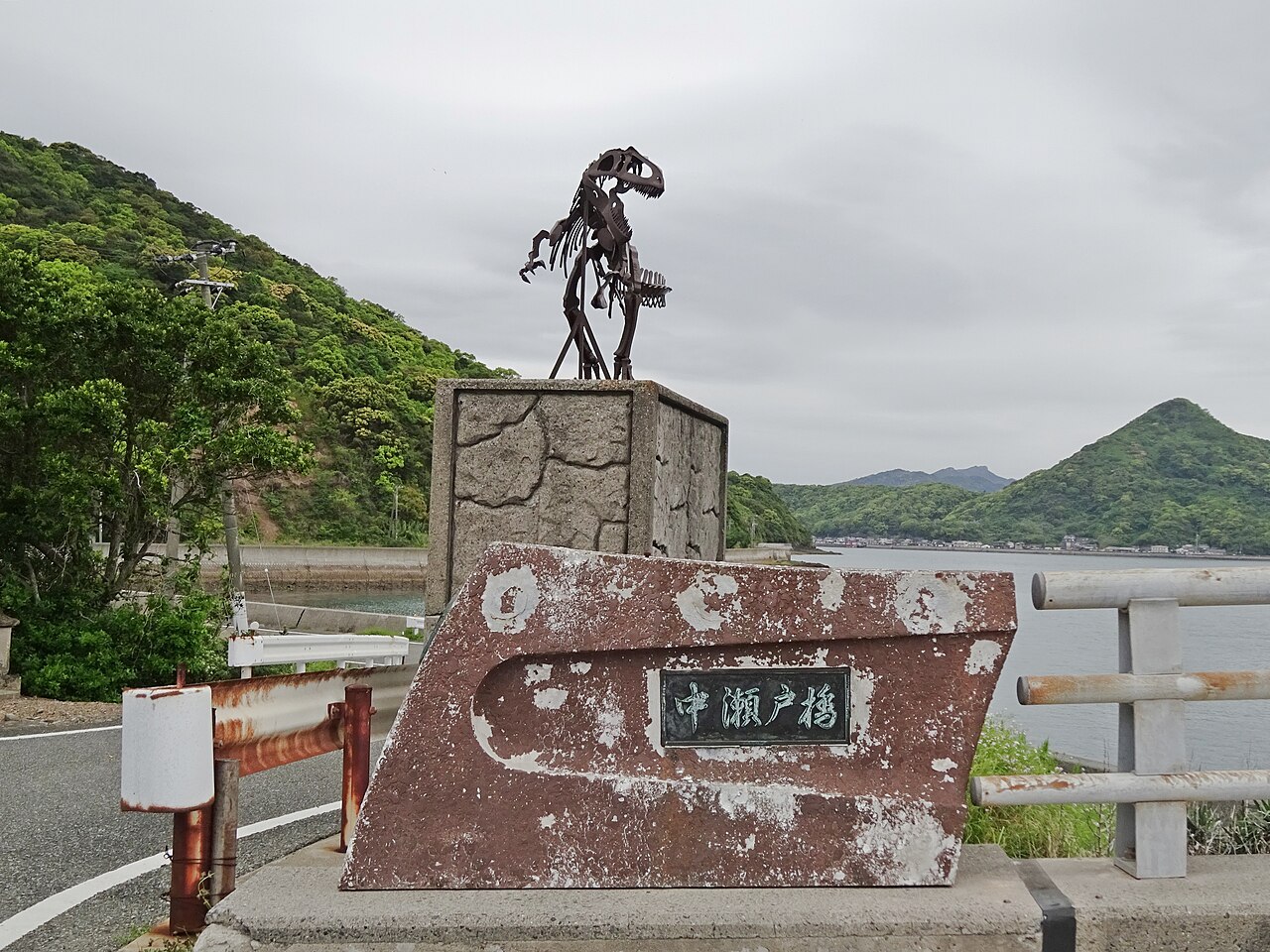 A dinosaur skeleton statue stands on a stone pedestal beside a rusty sign reading Nakaseto Bridge, with green hills and water behind.