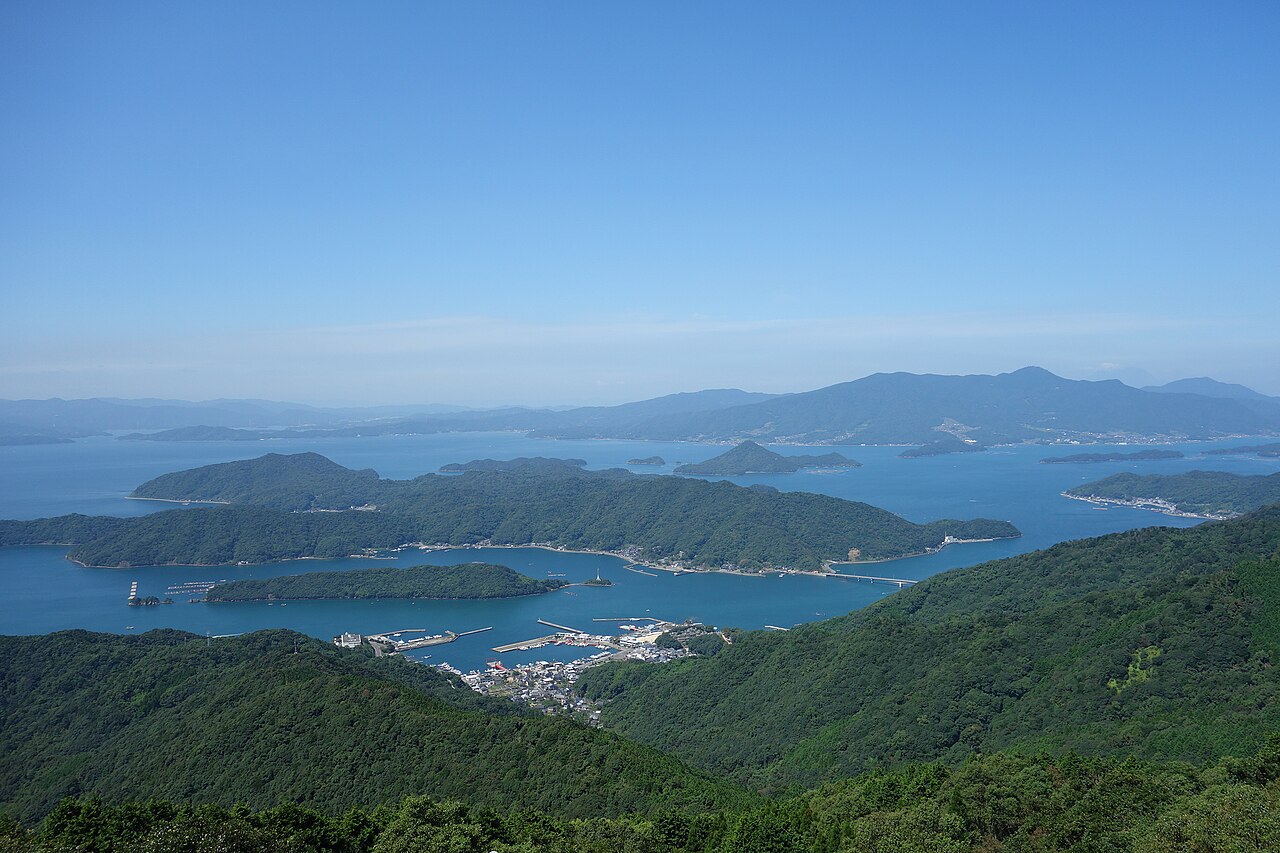 A wide view over blue sea and many green islands, with the small harbor town of Goshoura on the shore below.