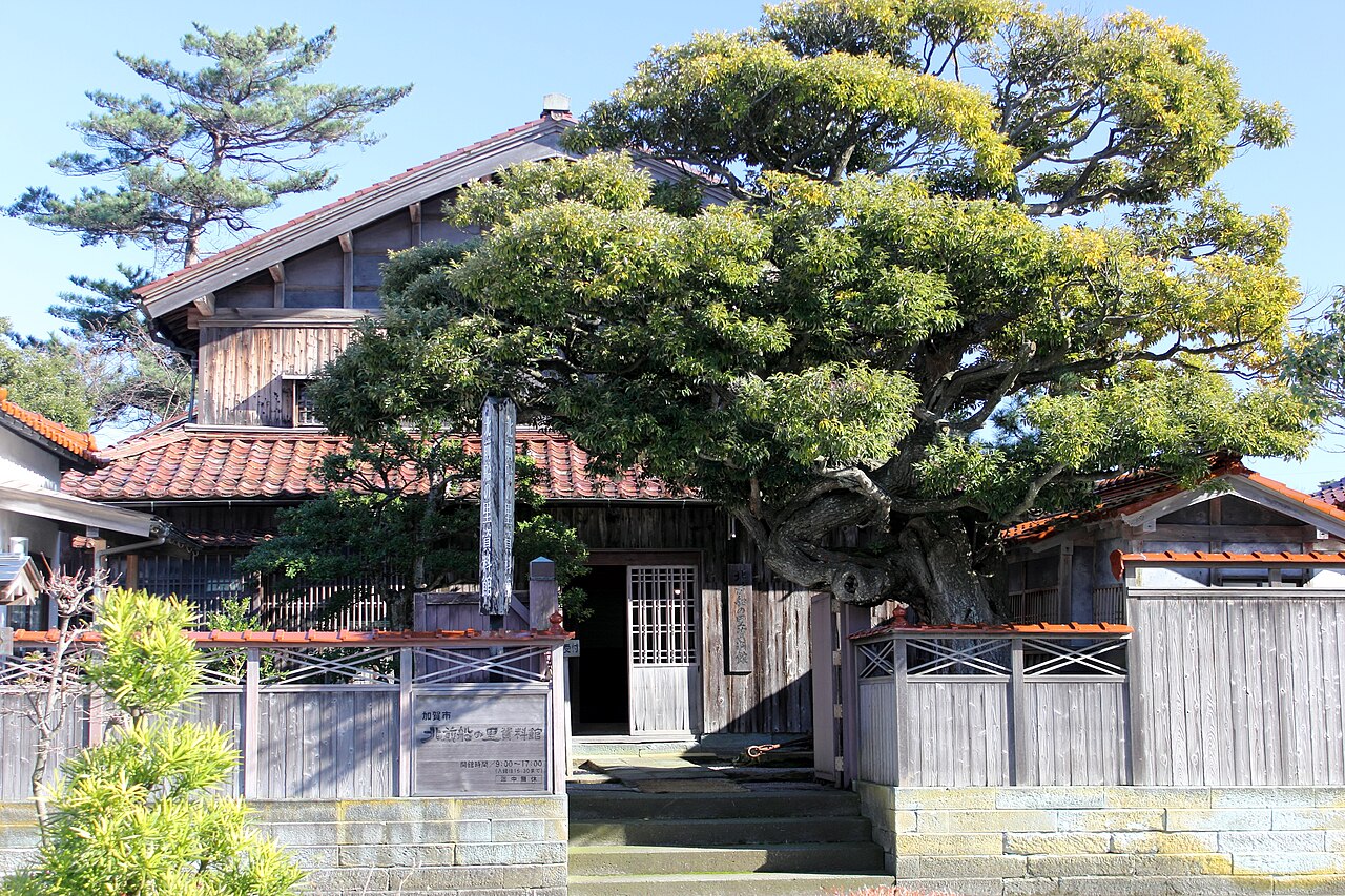 The Kitamaebune no Sato Museum in Hashitate: a wooden house with red tile roofs behind a large pine and a low board fence