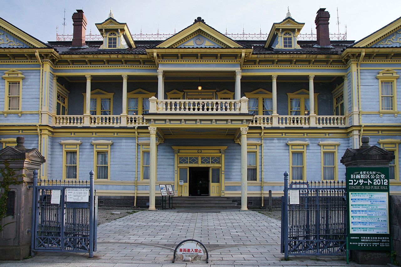 Front facade of the Former Public Hall of Hakodate Ward, a wooden Western-style building painted pale blue with yellow trim, a columned veranda with a balustrade above the entrance, iron gates on both sides and a paved approach in front