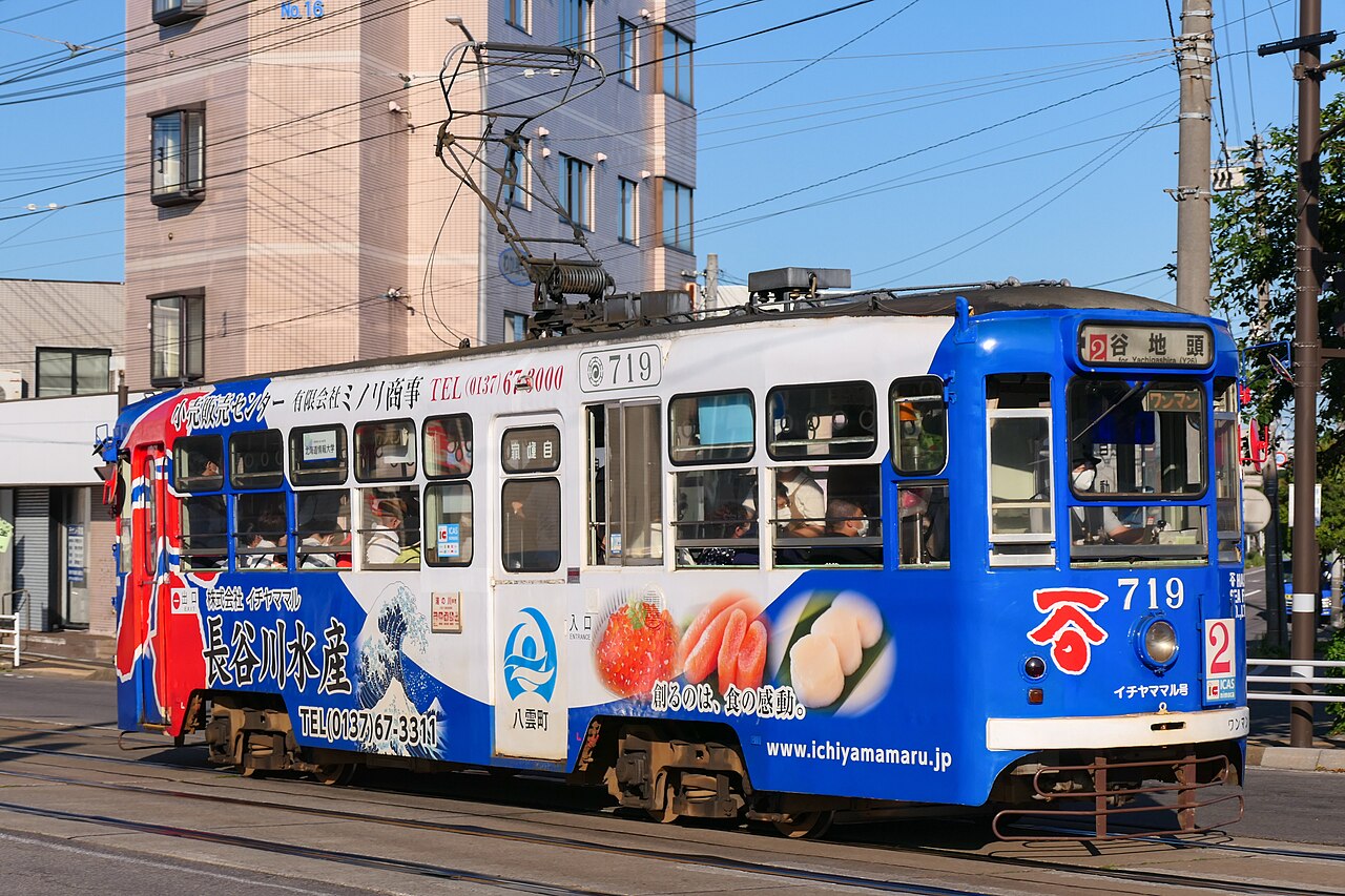 A blue and white Hakodate city tram with a seafood advertisement wrapped along its side, running on the street in daylight beneath overhead wires, with a pale apartment building behind