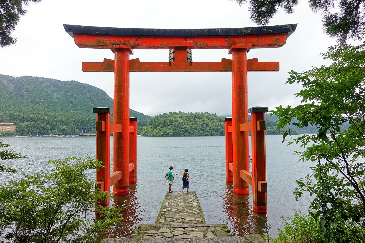 A tall vermilion torii standing in the shallows of Lake Ashi at the end of a short stone landing, with wooded hills across the water