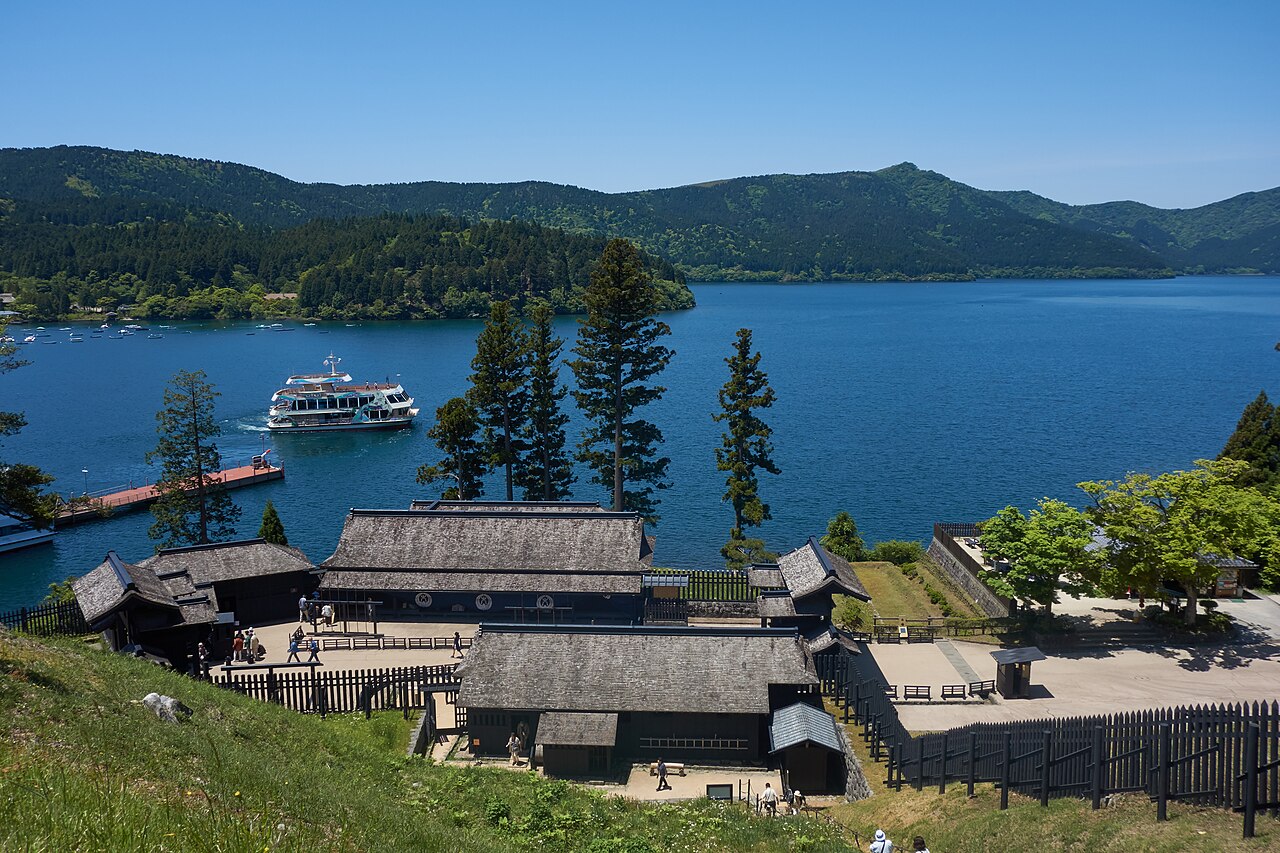 The dark timber buildings of the reconstructed Hakone checkpoint seen from above, with pine trees, a pleasure boat and the blue water of Lake Ashi behind