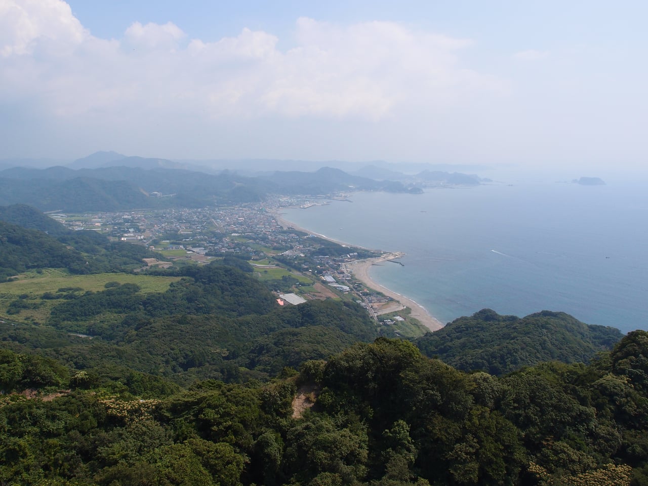 The view from Mount Nokogiri over wooded hills to a small coastal town and harbor on a calm blue sea.