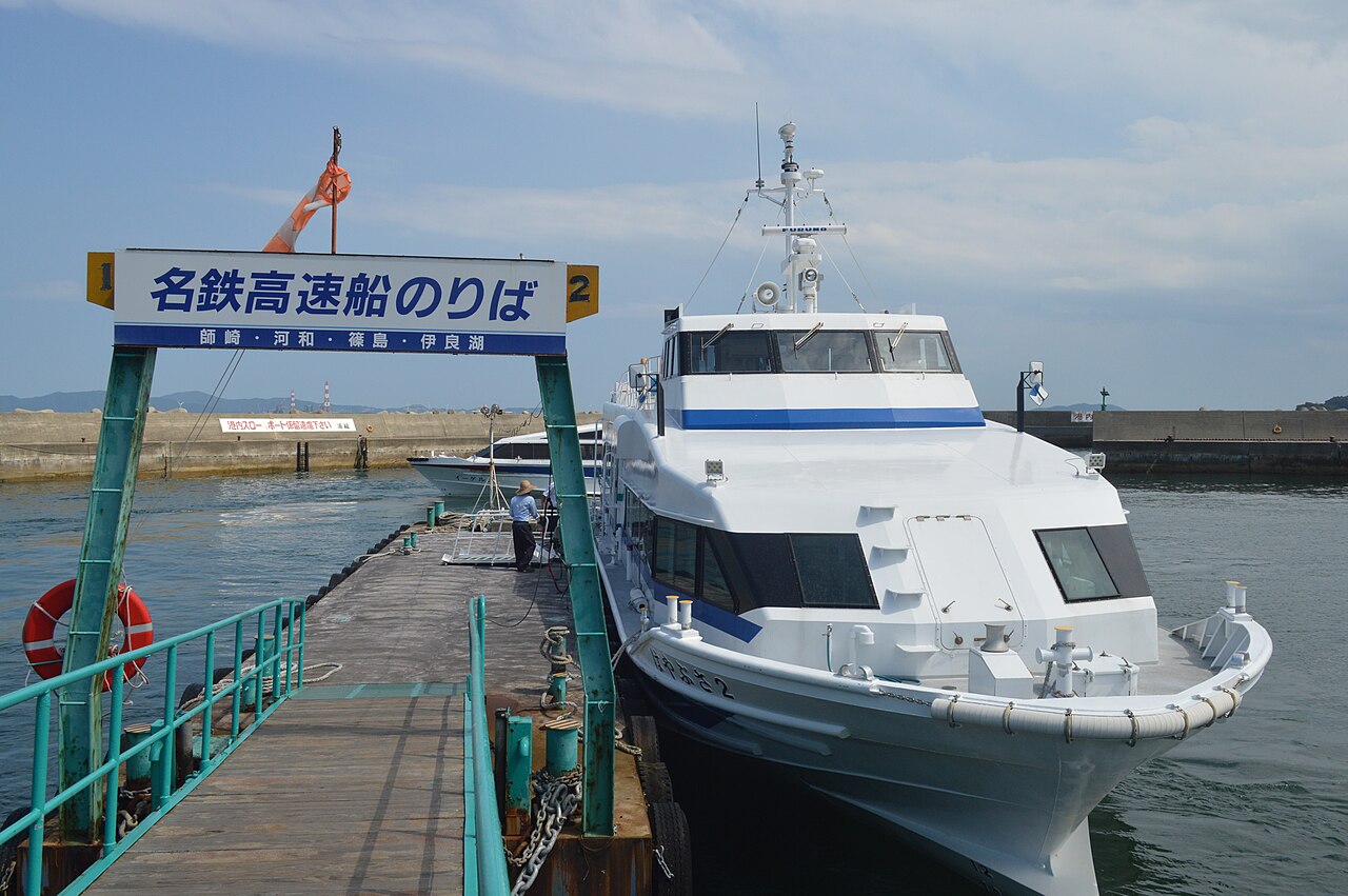 A white Meitetsu high-speed boat moored beside the boarding pier at Himakajima East Port, under a sign listing Morozaki, Kowa, Shinojima and Irago.