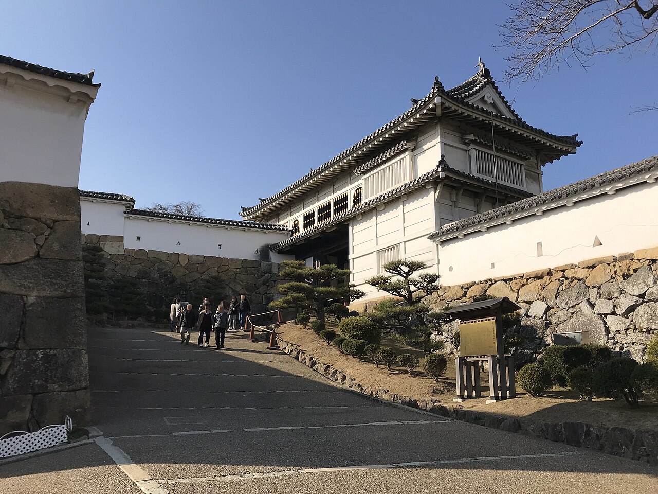 A gate tower with white plastered walls and a dark tiled roof above a stone base, seen from a sloping paved path with pine trees and a few visitors.