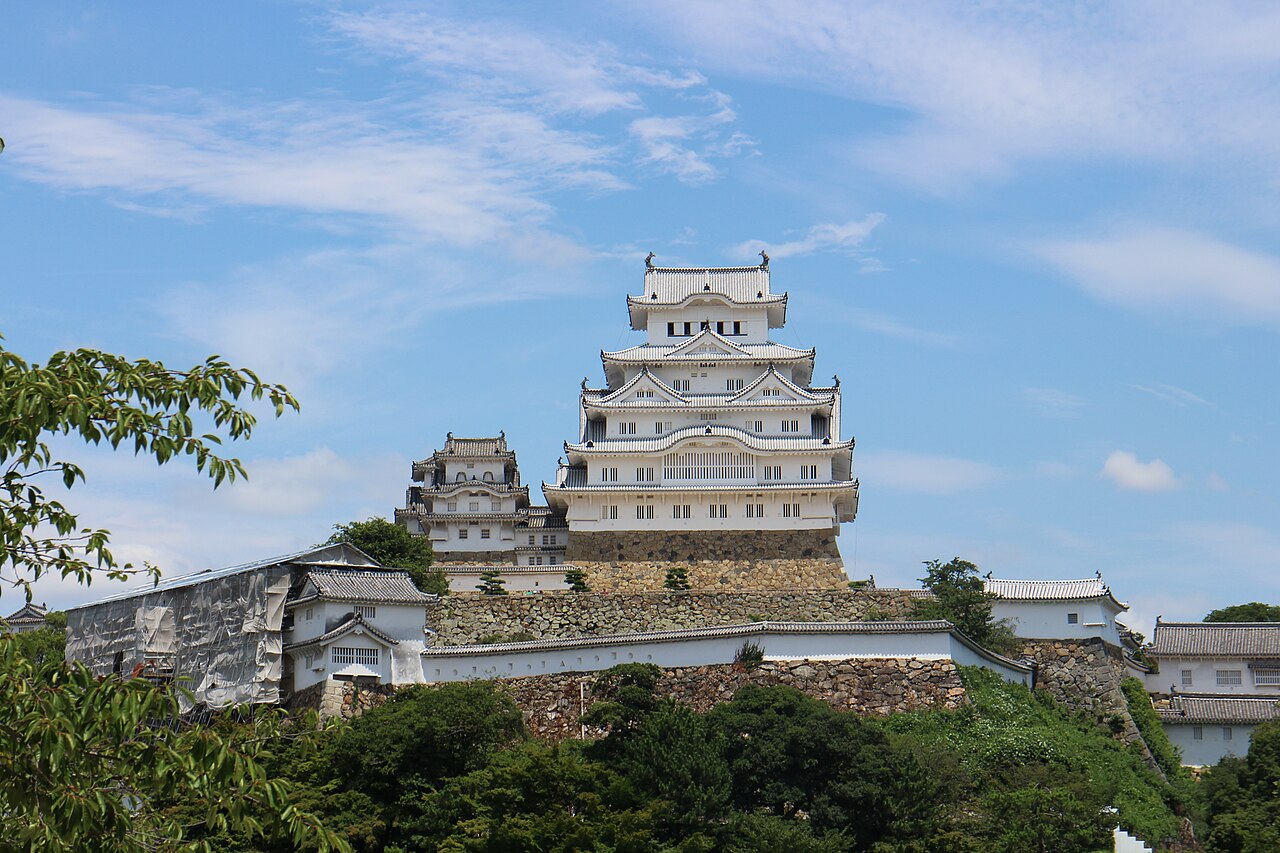 The white keep of Himeji Castle with stacked gabled roofs rising above a stone-walled hillside, with smaller turrets at left and trees in the foreground.