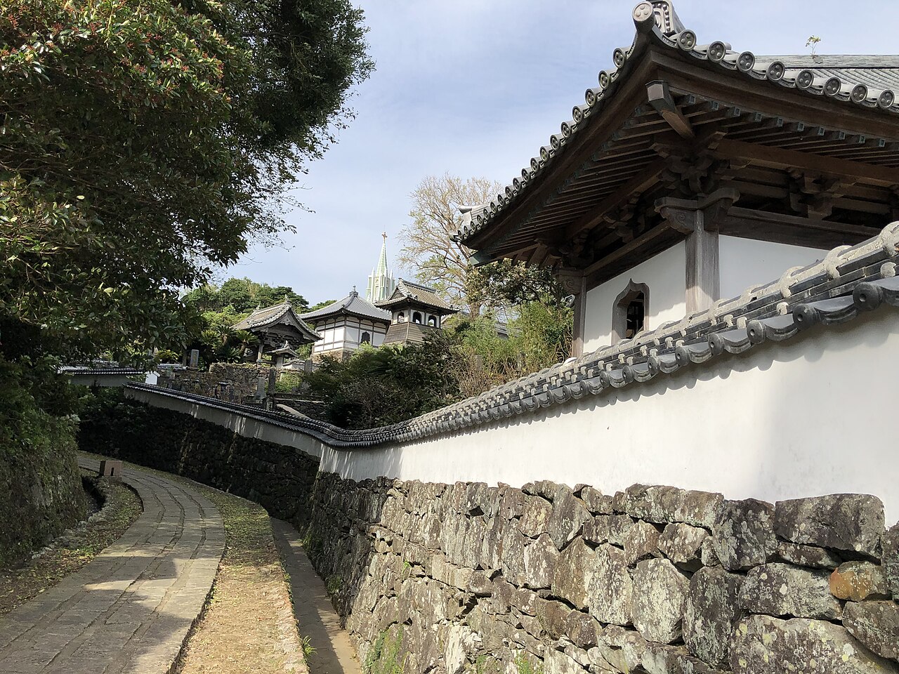 A stone-paved lane beside a white plaster wall, with temple roofs and a church spire beyond, in Hirado